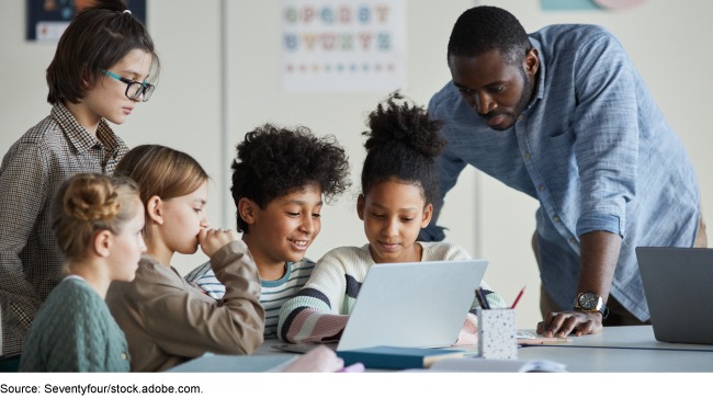 Five children gathered around a laptop with an adult looking over one of their shoulders.