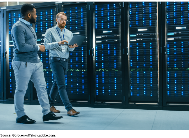 Two men, one carrying a laptop, walking in a server room