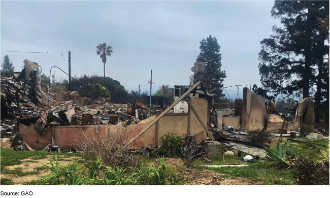 Partial burnt remains of a house’s walls and frames in the aftermath of a fire. Large piles of rubble are in the background. 