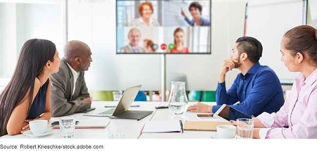 People working at table in a conference room with other people displayed on a videoconference screen.