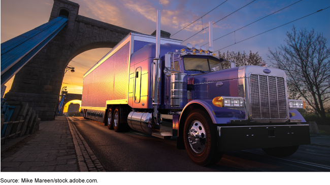 A tractor trailer driving under the arch of a bridge in low light.