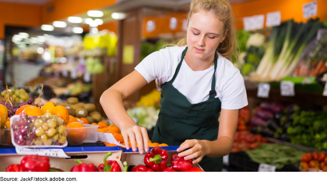 Teenage grocery worker wearing an apron, near boxes of red peppers.