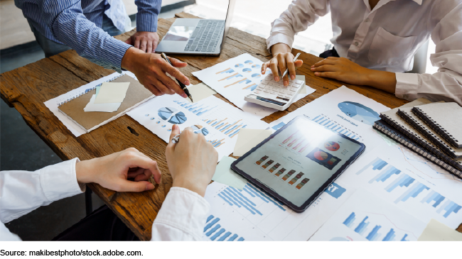 Three people sitting at a table with papers, a calculator, a tablet, and a laptop, many of which are showing data graphs