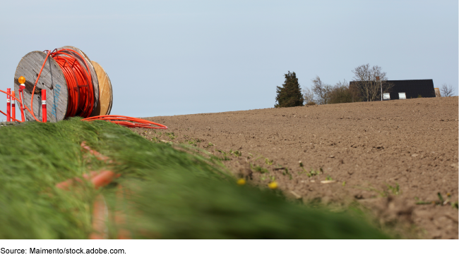 A plowed field with a building in the distance. On the left in the foreground is a giant spool of red cable.