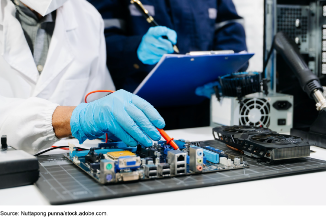 A person in a white coat and blue latex gloves is pointing to something on a computer motherboard with a tool while a person in the background holds a pen to a clipboard.