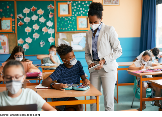 A teacher uses a tablet to show something to a young student at a desk.