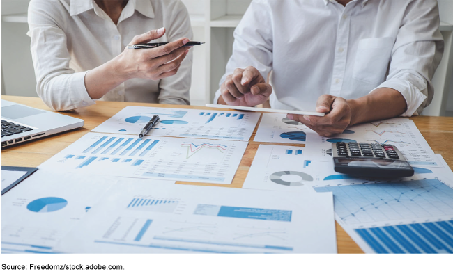 Two people at a desk with a laptop, a calculator, and papers with graphs and charts