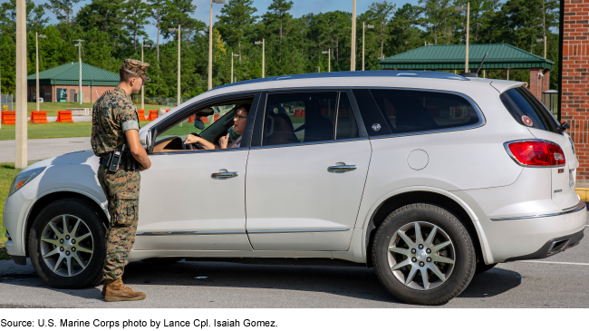 Service member in camouflage uniform speaks to the driver of a vehicle at a military checkpoint.