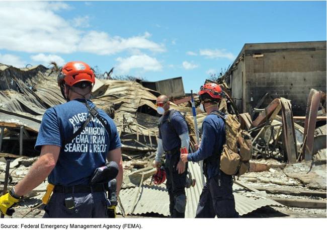 Three men wearing or carrying hard hats, masks, gloves, packs, and utility belts stand near the remains of a destroyed building. Three men wearing or carrying hard hats, masks, gloves, packs, and utility belts stand near the remains of a destroyed building.