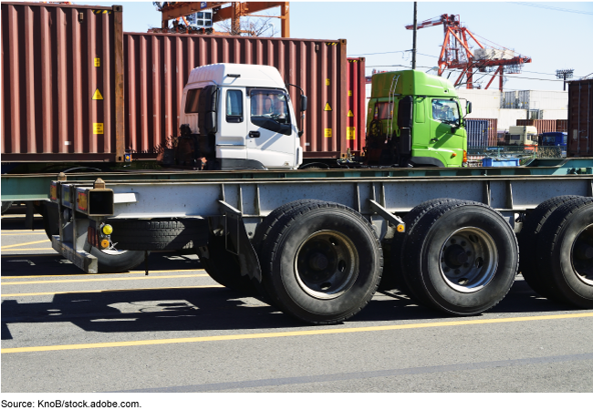 Semi trailer chassis with trucks, containers, and a crane in the background.