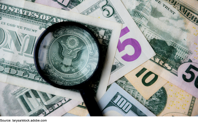 A pile of U.S. paper money of different denominations with a magnifying glass over the great seal on the back of the $1.
