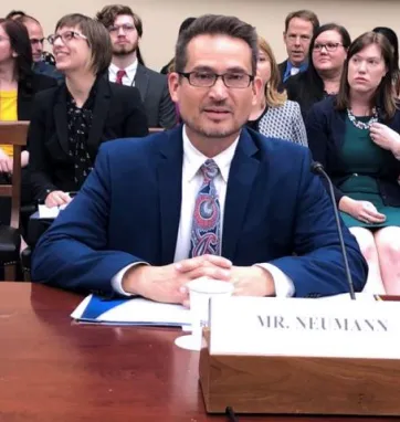 John Neumann testifying before congress is pictured sitting at a table speaking into a microphone