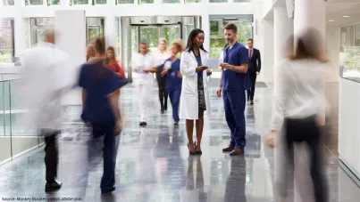 Image shows two doctors having a discussion in the midst of a busy hospital hallway as other doctors, nurses, and healthcare professionals walk past 