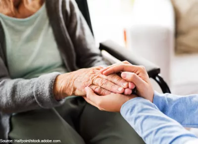 An image showing two people holding hands. One is older and seated in a wheelchair. The other is younger.