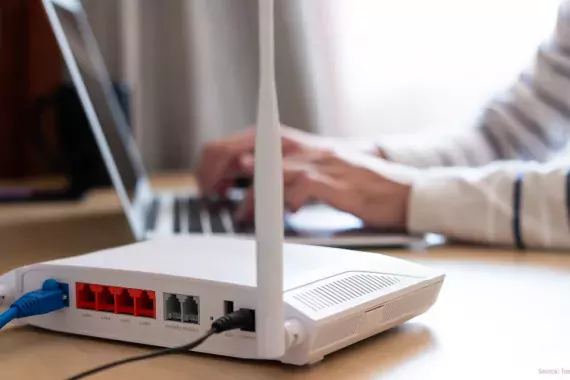 Photo showing a wifi router in the foreground and a woman at a laptop typing in the background