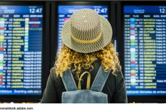 Photo showing a woman in a hat standing in front of an arrivals and departures board in an airport