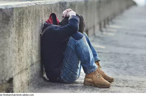 Stock image showing a little boy sitting on the side of the road with his arms over his head and knees to his chin.