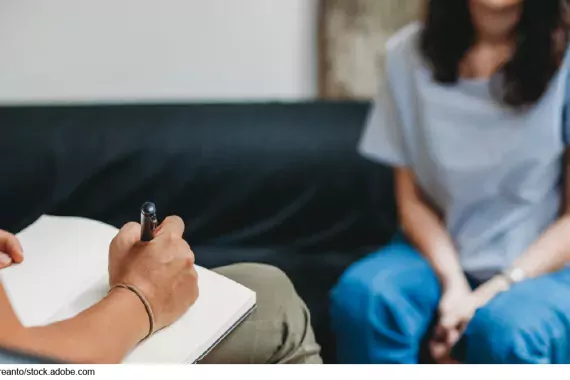 Stock image showing two people sitting across from each other. Nearest to us we see the arm and lap of a person with a clipboard taking notes. In the background, we see the neck-down of a person seated with their hands clasped.