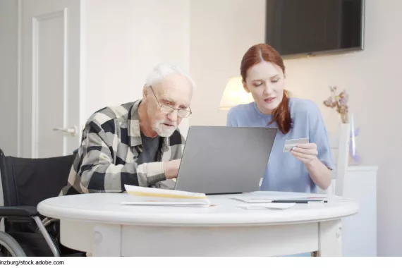 Photo showing an older adult sitting in a wheel chair next to a younger person. They are both working on a laptop together.