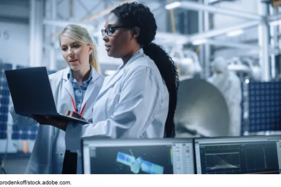 Researchers standing in a lab, surrounded by electrical and other equipment, looking at a laptop.