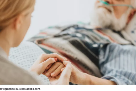 A woman holding the hand of an older man who is receiving supplemental oxygen from a medical device while in bed.