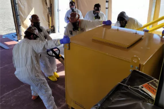 Workers in clean-suits and gas masks pull at a large, heavy-looking steel container