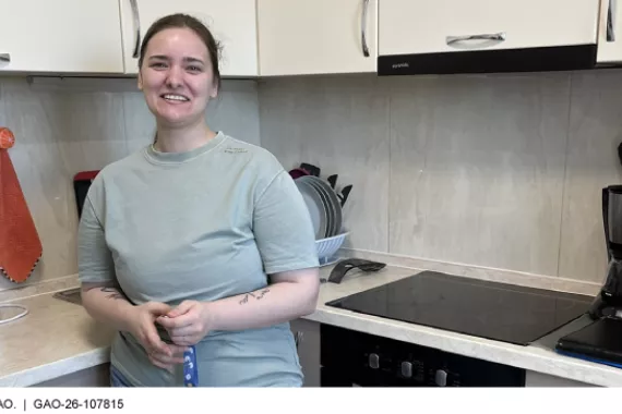 A person in a corner of an apartment kitchen, standing in front of a dishrack and next to a glass stovetop, with countertop appliances on either side 
