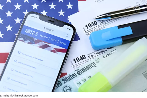 Tax documents, pens, and an iPhone on the official IRS website on a background of the U.S. flag.