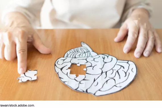 An older person's hands rest on a wooden table near a paper brain. The person rests a finger on a jigsaw puzzle-style piece of the paper brain, which is missing from the paper brain and off to the side.