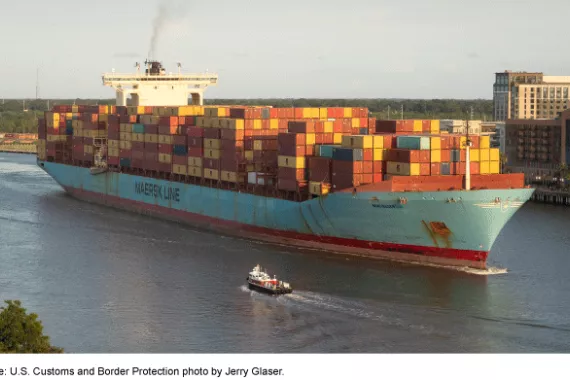 A large cargo ship transporting stacks of shipping containers on a body of water, with buildings and trees on the shoreline behind the ship.