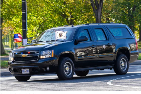 A dark colored SUV driving with two small flags on the front and blue and red lights in the front grill.