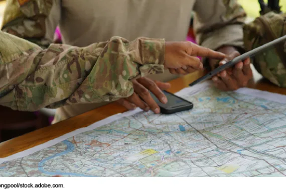 Two people in camouflage clothing pointing at a tablet while viewing a map and a phone on a table