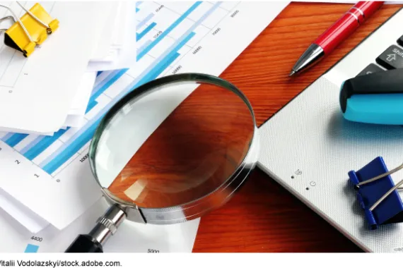 A magnifying glass lying on a desk surrounded by a laptop computer, papers, and pens.
