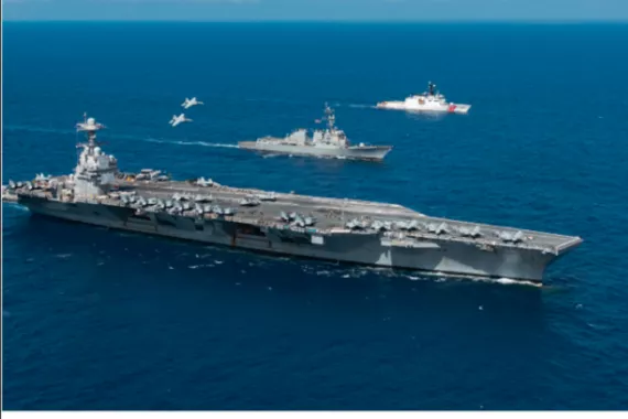 An aircraft carrier in the ocean with jets overhead and other ships nearby