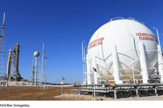 A landscape view of a large white tank that says liquified hydrogen, connected to a rocket ship and launch tower, with other tall metal structures nearby.
