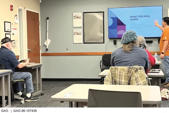 A classroom setting with older adults sitting at desks and a person pointing to a T.V. screen that says &amp;What you will learn.