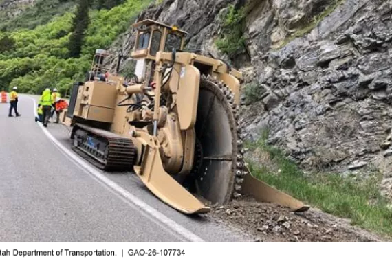 Several construction workers operate a larger trencher to excavate along the shoulder of a highway through a canyon.