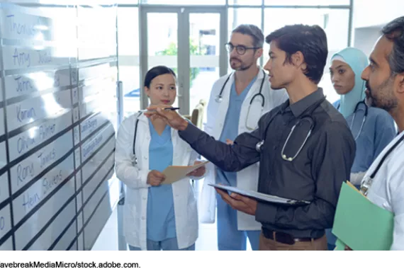 A group of people who all have stethoscopes draped around their necks look at a schedule written on a large whiteboard. Some are wearing scrubs, lab coats, or both. The person talking is wearing a dress shirt.
