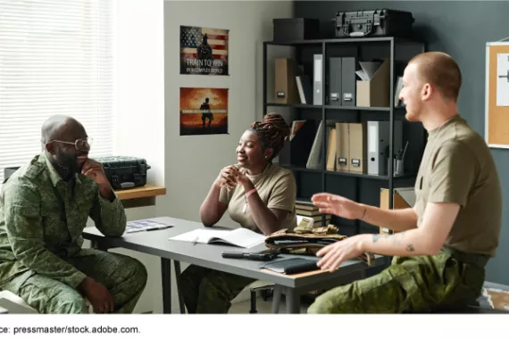 Instructor and cadets dressed in military attire socialize around a desk in a classroom. 