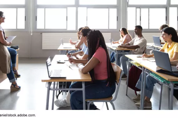 A classroom setting with young people sitting at desks looking at an adult holding a piece of paper at the front of the room.