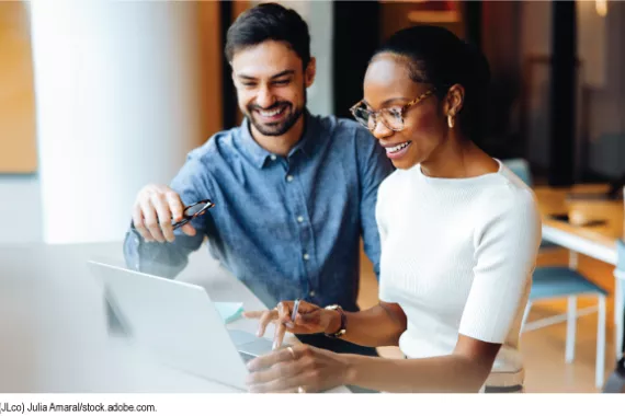 Two people at a desk using a laptop in a bright office setting. 