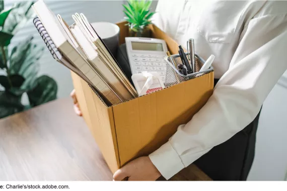 A person wearing a button-down shirt stands in an office, holding a cardboard box filled with various office supplies and a small plant.