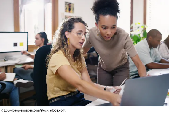 A woman sitting at a desk with a laptop in front of her while another woman stands next to her also looking at the laptop.