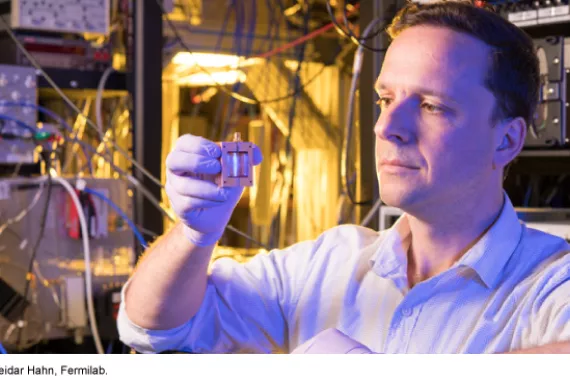 A man in a white shirt and rubber gloves holds a small box with internal circuitry in a room with complex electronic equipment.