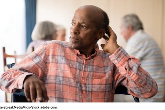 An older person sitting in a chair in a room with other older people in the background.