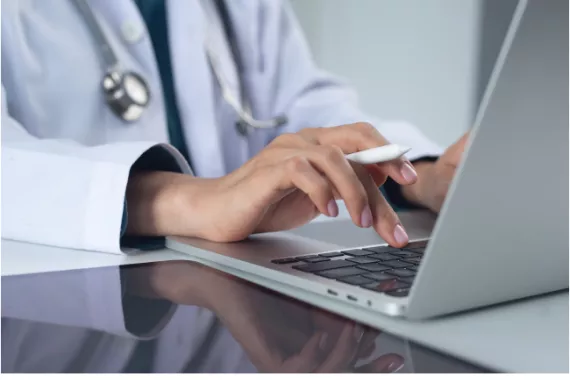  A person wearing a white coat and a stethoscope sitting at a desk typing on a laptop.