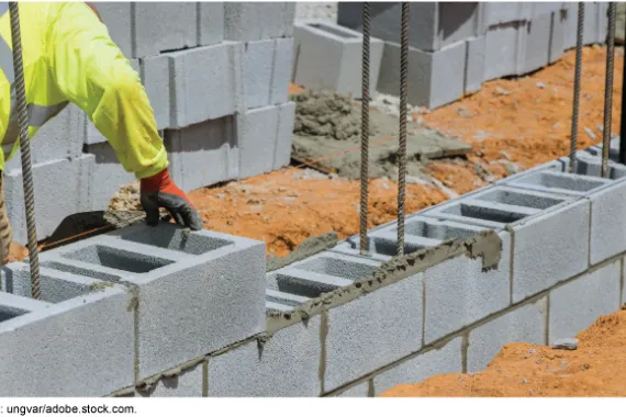 A mason builds a wall by laying a grey, precast concrete block on a mortared row.