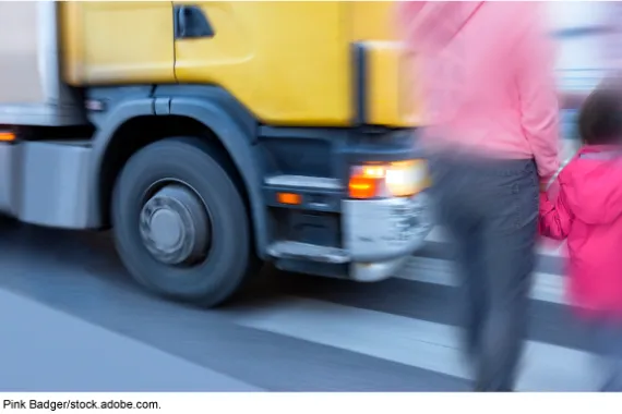 An adult holding the hand of a young child walking on a pedestrian crossing with a large vehicle closely approaching in front of them.