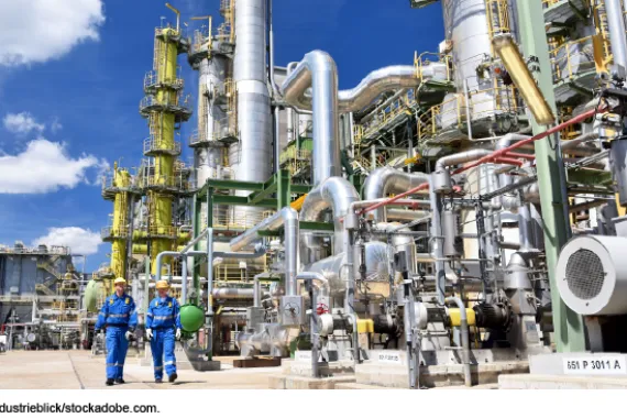 Chemical facility with pipes and two workers in blue jumpsuits and yellow hard hats.