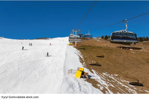 Chair lifts go up and down a steep hillside with brown grass below one side of the lift and groomed snow with skiers below the other.